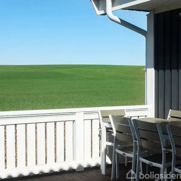 Terrassen har et sæt udendørsmøbler mod et grønne græsareal under en klar blå himmel. Hvide trægelænder omkranser området ved siden af et hus med mørke vægge.