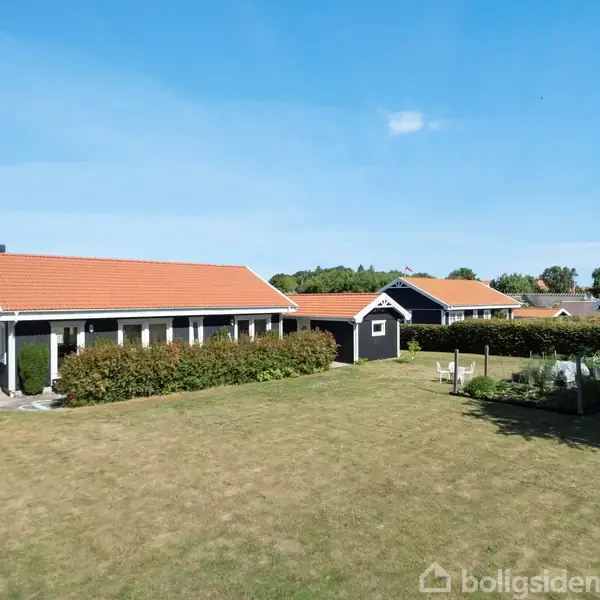 Et sort-hvidt hus med rødt tag står i en stor, solrig have omgivet af marker og træer. Flere sekundære bygninger kan ses i baggrunden.