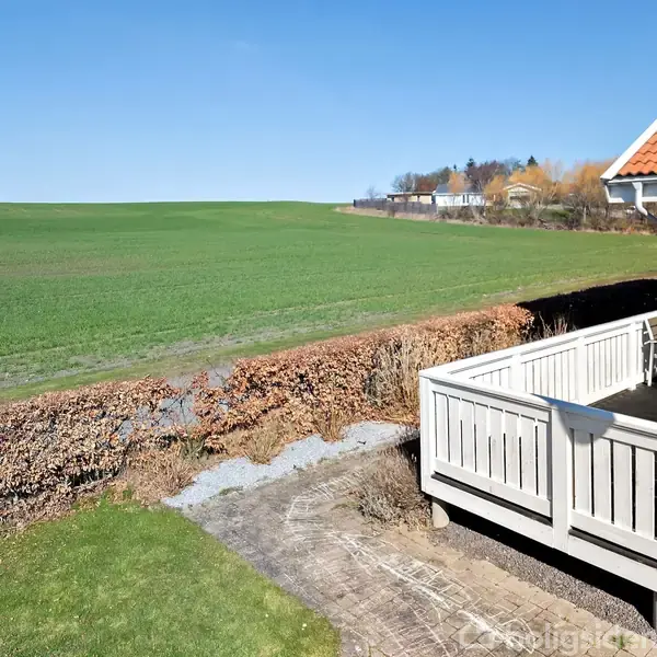Veranda med hvidt rækværk har et bord og stole, solbeskinnet. Omkringliggende grøn mark strækker sig, mens en markvej og buskads markerer adskillelse fra aftagende beboelsestræer i baggrunden.