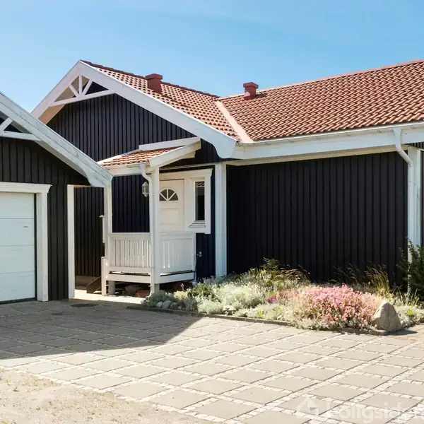 Sort, træbeklædt hus med hvide vinduer og rødt tegltag. Foran huset er en flisebelagt indkørsel, omkranset af blomsterbede og en garage med hvid port. Klar, blå himmel.