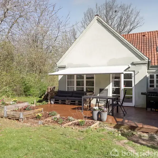 Huset har en terrasse med møbler og grill. Terrassen er omkranset af frodige buske og træer, og en grøn græsplæne strækker sig foran huset.