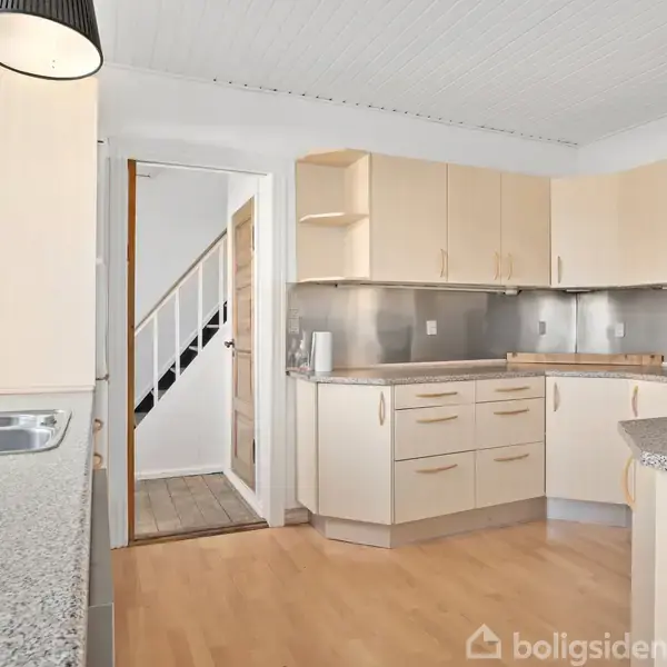 Et lyst køkken med lyse træskabe og sorte bordplader. En ovn er indbygget i skabene. Til venstre er en stålvask. En dør fører til en trappe.