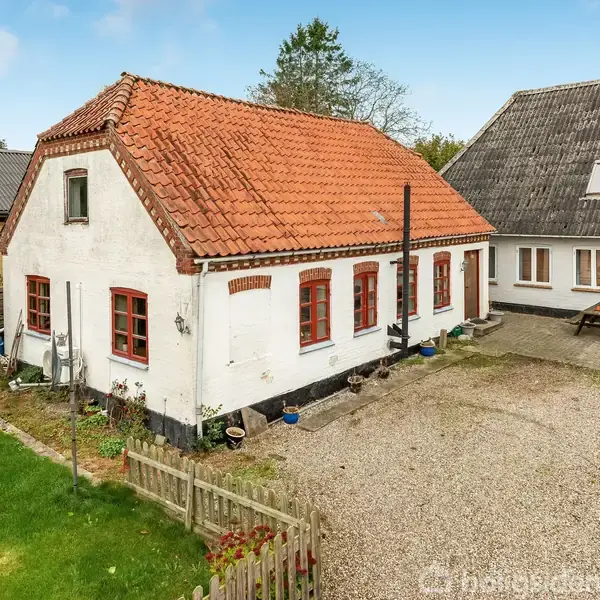 Et hvidt stuehus med rødt tegltag står stille foran en grusbelagt gårdsplads; til venstre ses en grøn have med hegn. Den himmel er klar med spredte skyer.