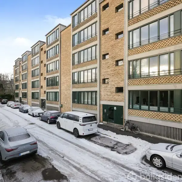 Flere etagebygninger med gule mursten står langs en sneklædt gade. Biler er parkeret i siden af vejen, og cykler står tæt på indgangen.