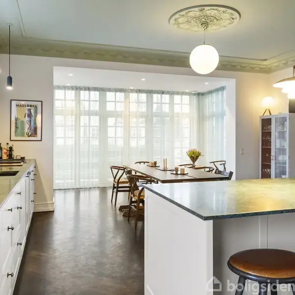 Køkken med hvide skabe og en stor, mørk spisebord i baggrunden. Moderne lamper hænger fra loftet med detaljeret stuk. Store vinduer giver naturligt lys. Plakat på væggen.