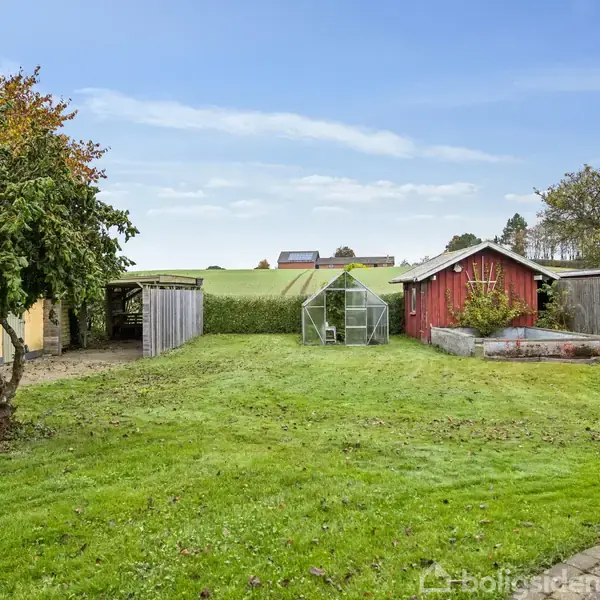 En velplejet græsplæne strækker sig mod et grønt drivhus og en rød skurbygning. Træer og en flagstang omkranser plænen, mens en himmel med lette skyer ses i baggrunden.