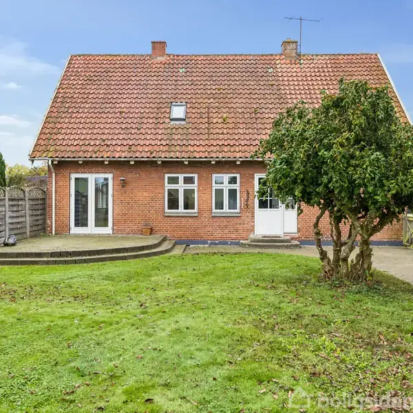 Et rødt murstenshus med rødtag står i en velplejet have med en græsplæne. En terrasse af træ ligger foran huset, hvilket skaber en hyggelig atmosfære.
