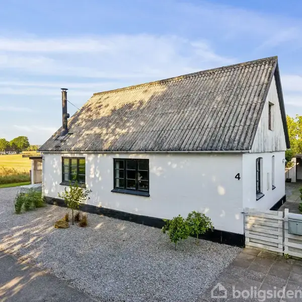 Huset står stille på en grusvej omgivet af grønne marker og træer under en klar blå himmel. Husnummeret "4" er synligt på væggen ved siden af døren.