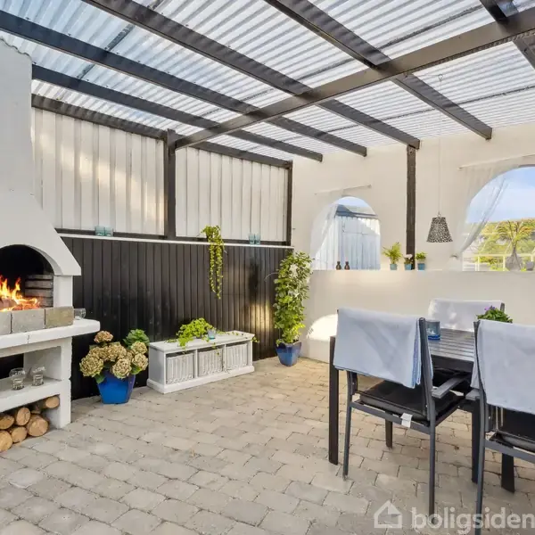 Pejs med ild brænder under et halvtag i en udendørs terrasse. Omkring er der stole med hvide betræk ved et bord og planter i krukker.