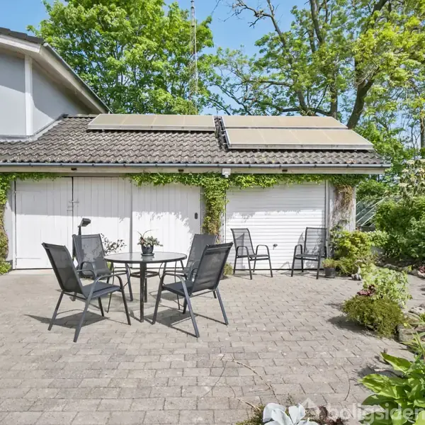 En terrasse med et rundt bord og fire stole står foran en garage med hvide døre. Solpaneler ses på taget, og omgivelserne er grønne og frodige.
