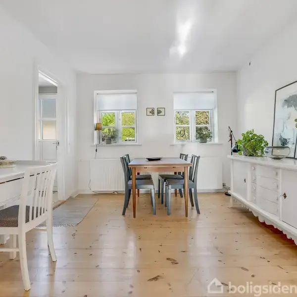 Et lyst rum indeholder et spisebord med stole. To vinduer slipper lys ind. Der er dekoreret med planter og billeder på væggene, og der er en skænk og et skrivebord.