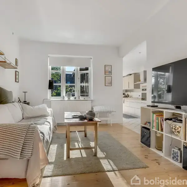 En lys stue med en hvid sofa til venstre, en træbord til højre, og et stort TV på en reol. Bagved er et vindue, og der er udsigt til en grøn have.