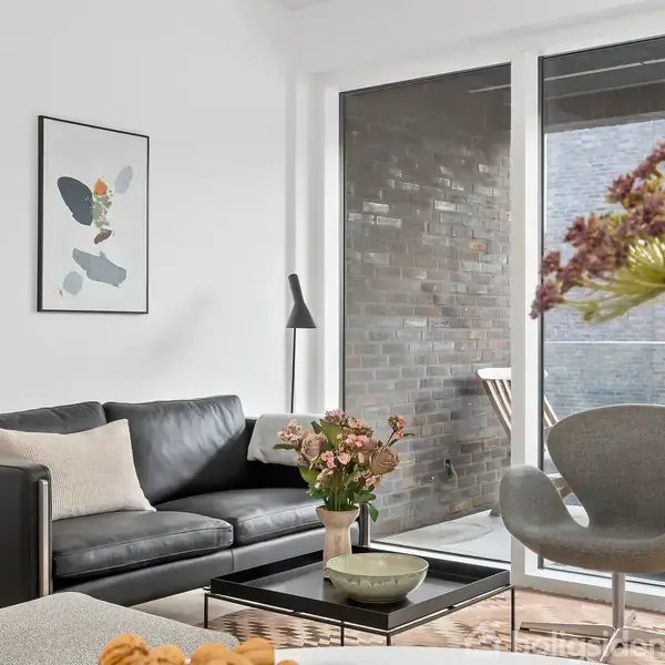 En moderne stue indeholder en sort lædersofa med hvide puder, placeret ved et sofabord med blomster. Store vinduer giver udsigt til en murstensvæg. En plakat hænger på væggen.