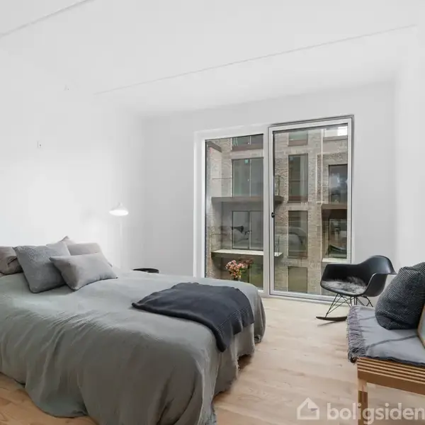 Soveværelse med en dobbeltseng dækket af gråt sengetøj, en stol ved en glasdør, der fører til en balkon. Et billede er lænet op ad væggen på en træbænk.