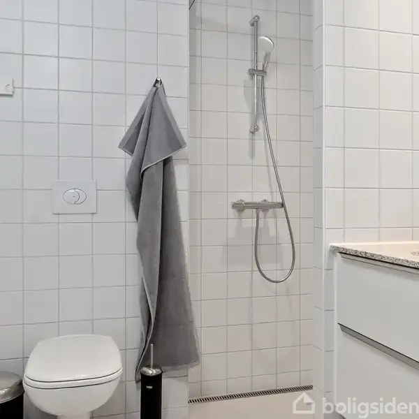 Toilet i moderne, hvidt flisebadeværelse; et gråt håndklæde hænger på væggen ved siden af en brusekabine med bruser. Der er et hvidt skab med vask til højre.