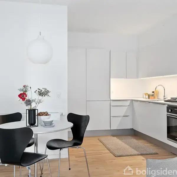 Et hvidt køkken med indbyggede apparater og træbordplader. Rundt spisebord med sorte stole og blomster i vase. Lysindfald fra minimalistisk lampe bidrager til den lyse atmosfære.