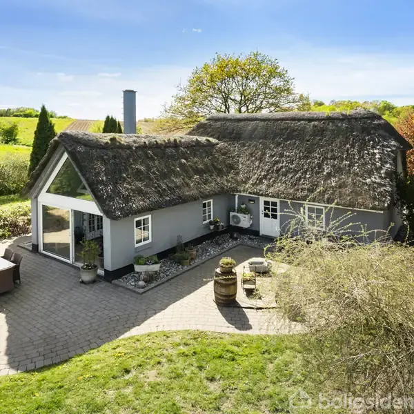 Stråtækt hus med græs- og buskomgivelser. Huset har store vinduer og en terrasse med havemøbler. Omgivet af frodig natur, herunder træer og velplejet have. Ingen synlig tekst bortset fra "Boligsiden".