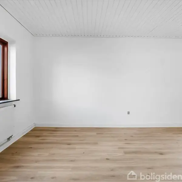 Et tomt rum med lyst trægulv, hvide vægge og loft. To store vinduer til venstre og højre giver naturligt lys. Radiatorer er placeret under vinduerne.
