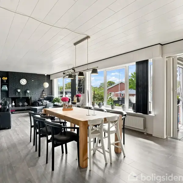 En spisestue indrettet med et langt træbord og stole, omgivet af en moderne stue med sofa, store vinduer der giver udsigt til have og altan med havedør åben.