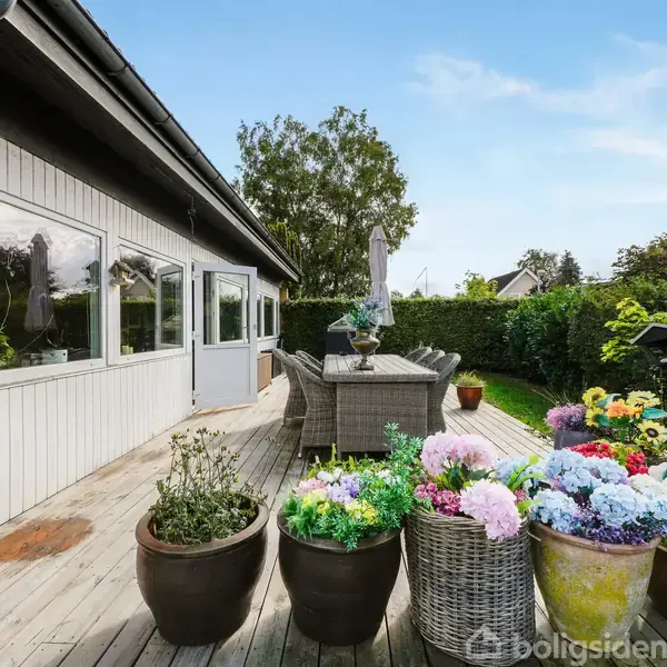 Terrasse med blomsterkrukker, bord og stole under en parasol. Omgivet af hække og træer, giver en hyggelig udendørs atmosfære på en solrig dag.