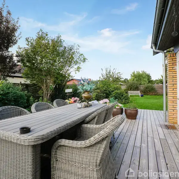 Terrassebord omgivet af flettede stole står på en træterrasse ved siden af et hus. Haven er fyldt med grønne buske og træer under en blå himmel i baggrunden.