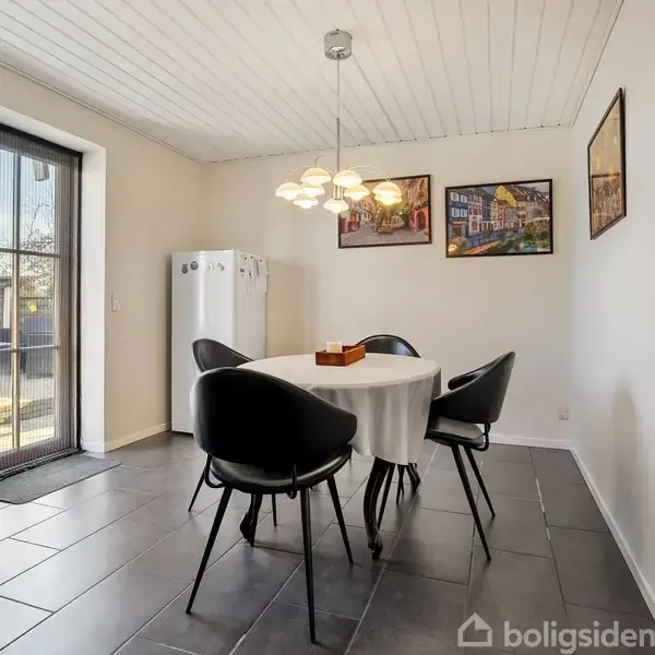 Rundt spisebord med hvid dug og fire sorte stole står i et lyst rum med hvide vægge. En pendellampe hænger over bordet. Rummet har flisegulv og en skydedør til haven.