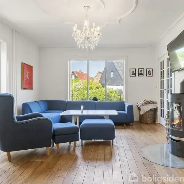 Stue med blå hjørnesofa og stol ved brændeovn. Et sofabord står foran vinduer med udsigt til grøn have og huse. TV hænger på væggen, og en lysekrone hænger i loftet.