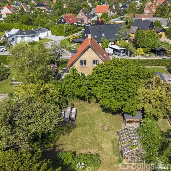 Et to-etagers hus med rødt tegltag er omgivet af frodige træer og buske. I haven står et havebord med stole på græsplænen. Opkørslen ses i baggrunden.
