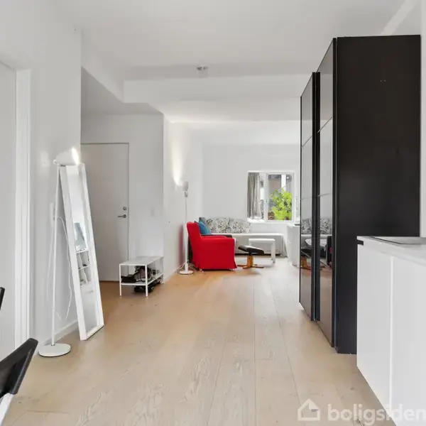Spejl står ved en væg i en moderne lejlighed, hvor en køkkenenhed er placeret langs højre væg. Stol og bord ved siden af. Rød sofa ses i baggrunden.