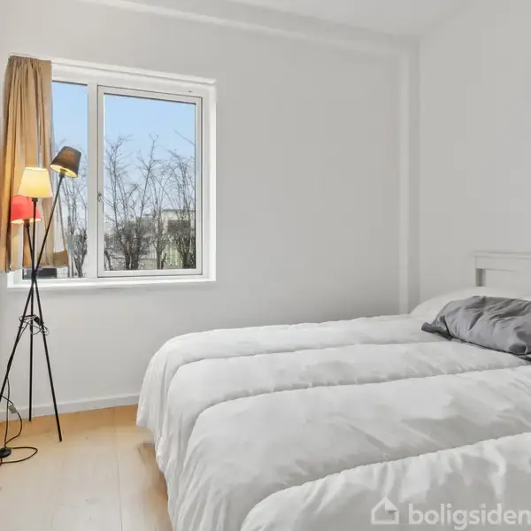 Seng med hvidt sengetøj står ved siden af et vindue. En gulvlampe i sort står til venstre. Udenfor ses bare træer. Rummet er lyst og minimalistisk.
