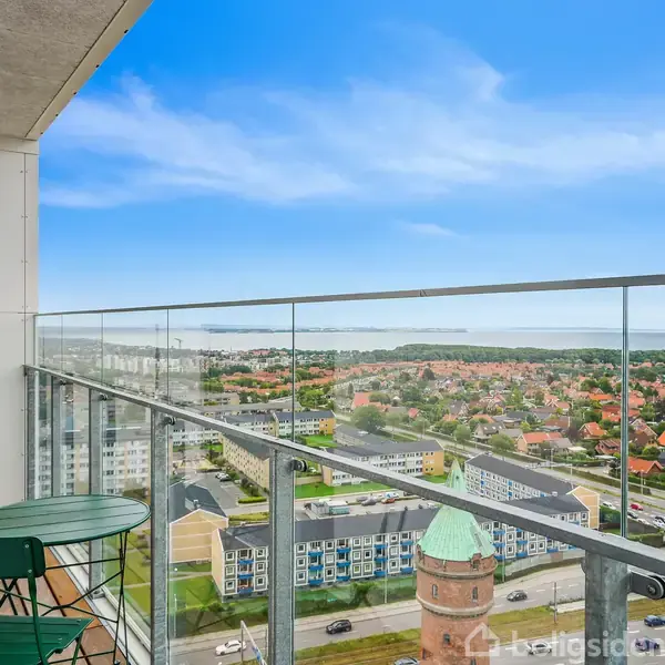 Balkon med grønne havemøbler set fra siden, med glasværn. Udsigt over en by med mange huse og grønne områder i baggrunden under en blå himmel.