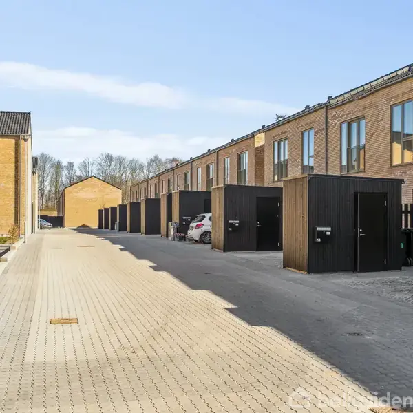 Rækkehuse står i en række langs en brolagt vej med parkerede biler ved siden af træskure. Området er omgivet af træer og en klar blå himmel uden skyer.