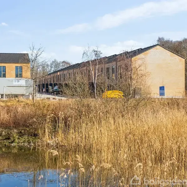 Flade tage på rækkehuse strækker sig langs en vej, omkranset af græs og træer, med en dam i forgrunden. Bygningerne er murstensfarvede med blå døre og vinduer.
