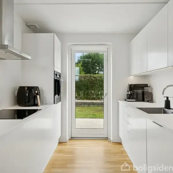 Et moderne køkken med hvide skabe og apparater, som kaffemaskine og ovn. En glasdør fører ud til en grøn have. Trægulv skaber en varm kontrast til det hvide interiør.