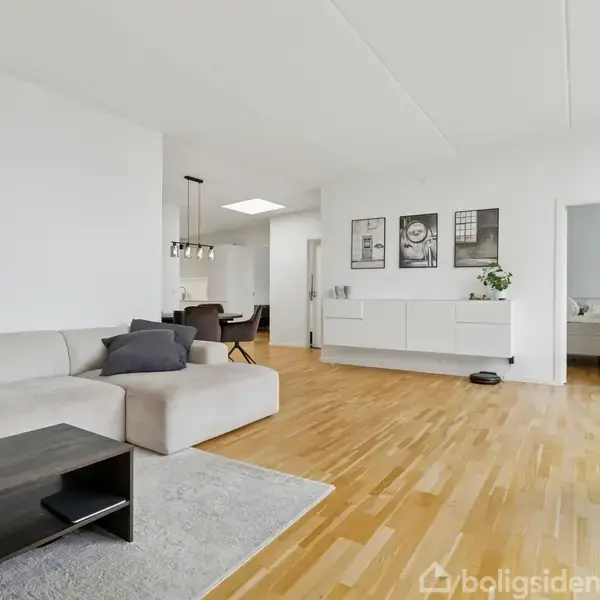 Sofa står i moderne stue med trægulv, hvorpå et sofabord og tæppe er placeret. Reol pryder væggen, og dør åbner til soveværelse. Grøn plante står i hjørnet.