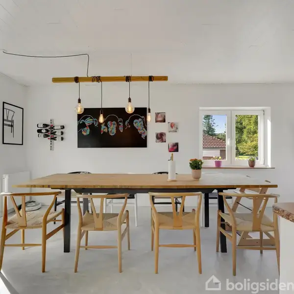 Et spisebord med seks stole står i et lyst rum. Tre hængelamper lyser over bordet. Væggen har kunst og et stort vindue, mens en dør fører ud til en terrasse.