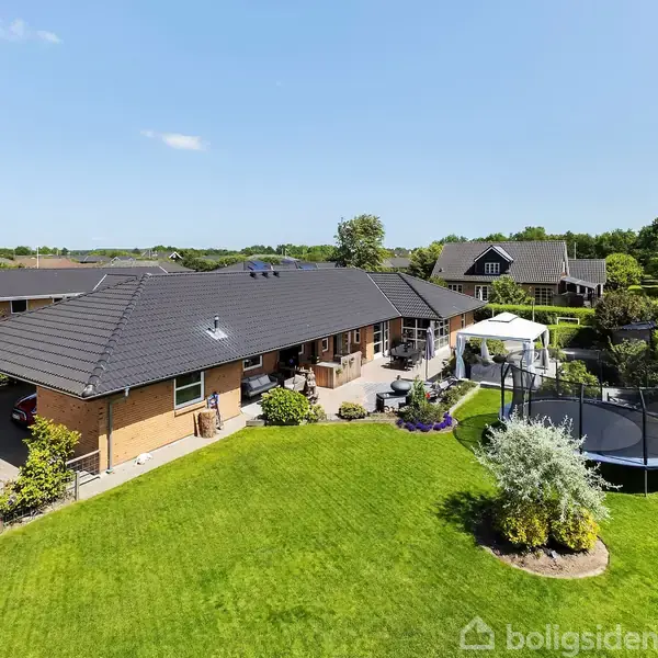 Et stort, moderne parcelhus med have og trampolin. Huset har en mørk tagkonstruktion og terrasseområde. Haven er omgivet af velholdt græsplæne og grønne buske, typisk for en forstad.