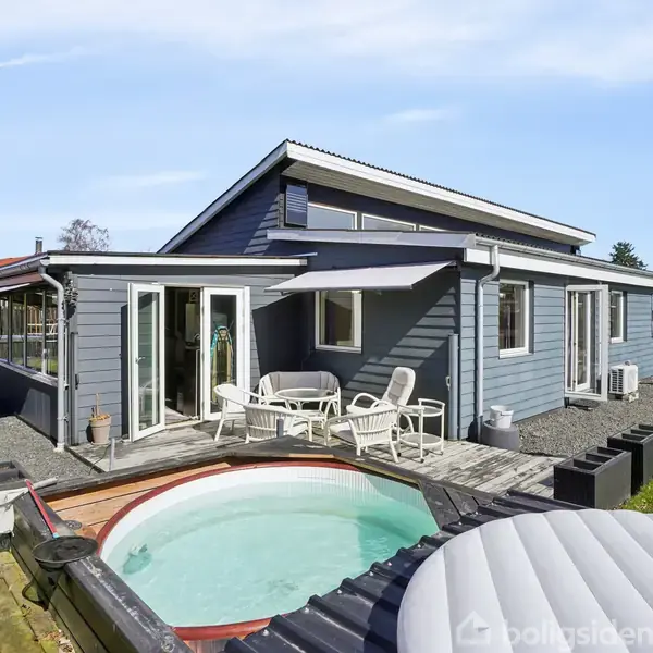 Blåt hus med terrasse og hvidt gelænder. Foran huset er der en rund pool ved siden af et overdækket opholdsområde med bord og stole. Græsplæne omgiver området.