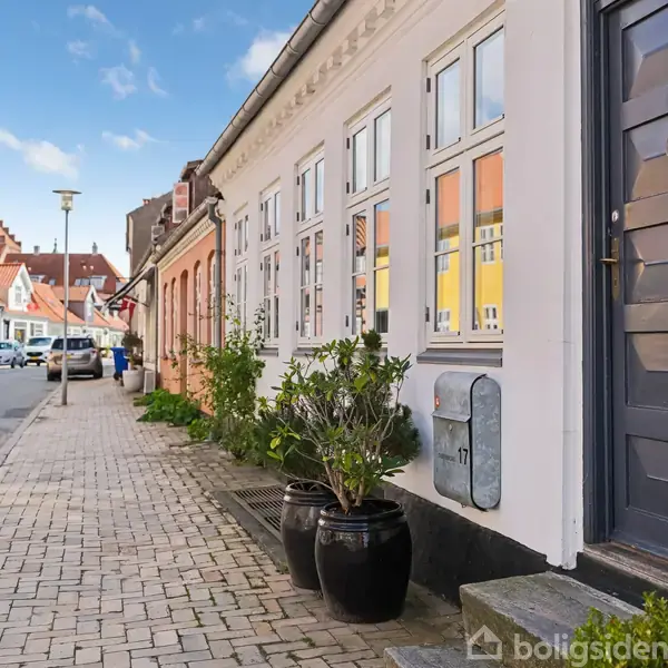 En række byhuse står langs en brostensbelagt gade med blå himmel i baggrunden. En sort dør med tallet "17" ses i forgrunden, omgivet af planter i krukker.