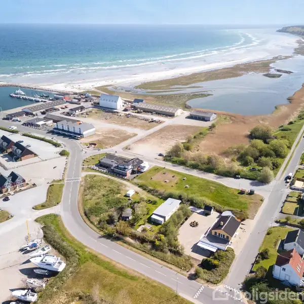 Kystområde med vindmøller ved havn, både i vandet, landevej og huse til venstre. Til højre ses en række huse langs en vej, med hav og strand i baggrunden.