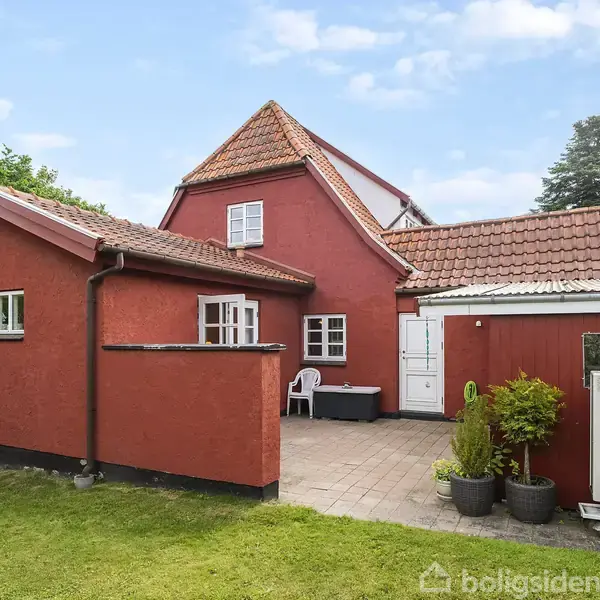 Et rødt hus står i en have. Det tilknyttede fliseområde har en dør, stole og planter. Området er grønt og fredeligt med træer i baggrunden under en blå himmel.