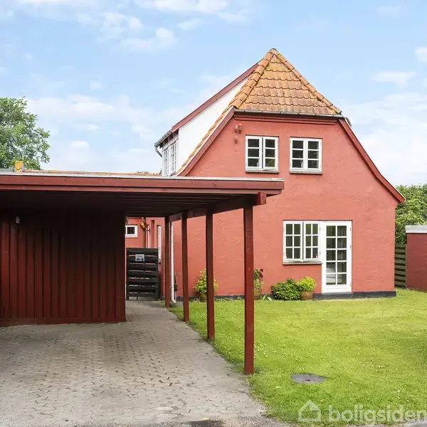Et rødt hus med hvide vinduer og rødt tegltag står i baggrunden. En carport står foran huset, omkranset af grønne hække og græs.