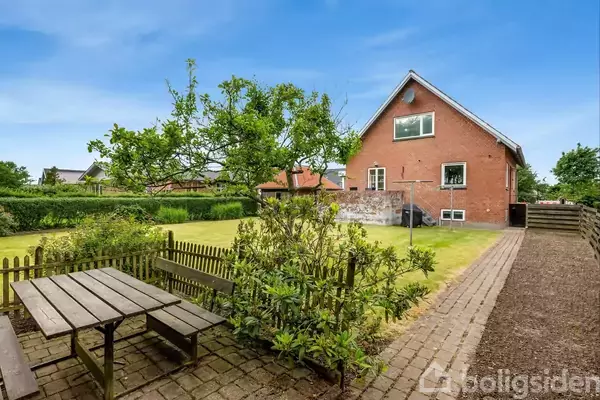 En rød murstensbygning står i en grøn have med en træterrasse. Ved siden af ses et picnicbord på en brolagt terrasse omgivet af buske og et lille hegn.