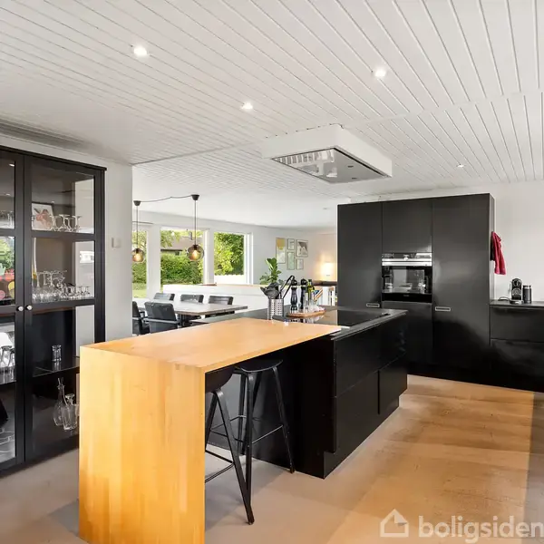 Et moderne køkken indeholder en sort køkkenø med barstole og indbyggede skabe. Til venstre står et vitrineskab med glas. Store vinduer giver udsigt til en lys stue med spisebord.