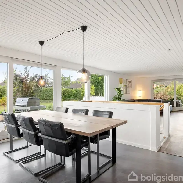 Spisebord med seks sorte stole står i en lys spisestue med store vinduer, der viser en have udenfor. Et åbent køkken er i baggrunden.