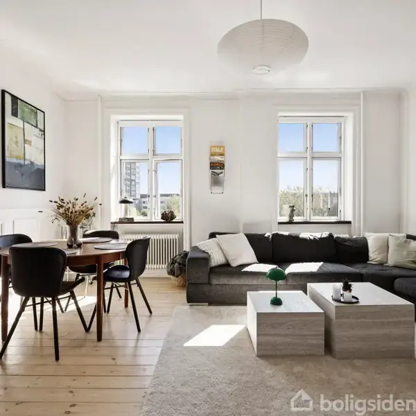 En stue med et stort mørkegråt hjørnesofaarrangement, står i et lyst rum med trægulv. Der er et rundt spisebord med sorte stole og moderne kunst på væggen. Tre små borde står foran sofaen. To store vinduer giver naturligt lys.