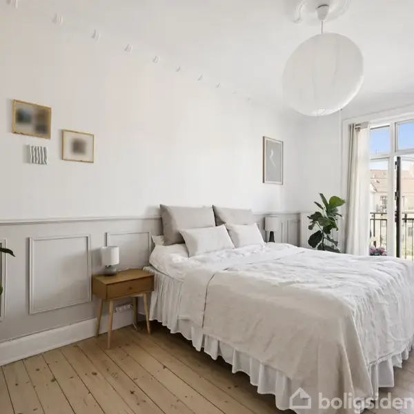 En seng med hvidt sengetøj står i et lyst soveværelse. Ved siden af er et lille natbord og grønne planter. Værelset har hvide vægge og en dør til en balkon.