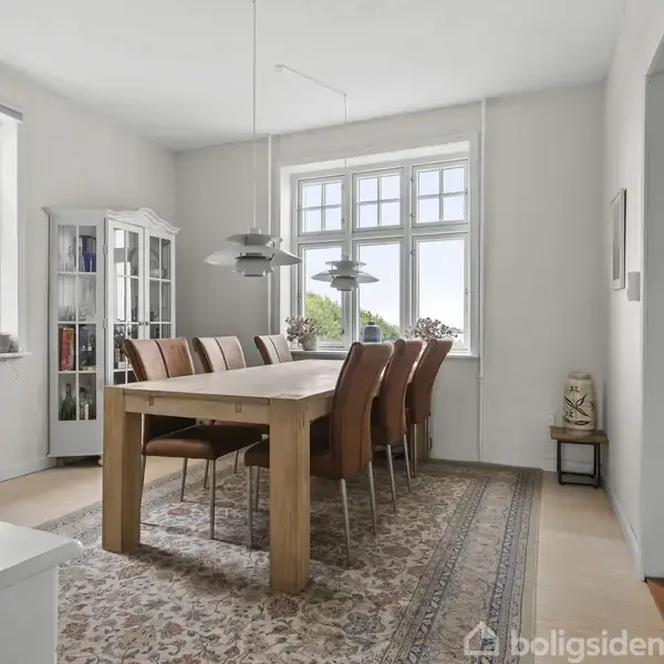 Et spisebord omgivet af otte polstrede stole står på et mønstret tæppe. Rummet har hvide vægge, store vinduer og en vitrine med bøger i hjørnet.
