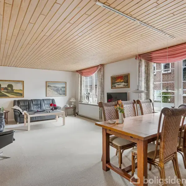 Stue med træloft og tæppebelagt gulv. Sofa og stole står mod væggen, og et spisebord med otte stole er i rummet. Store vinduer med gardiner slipper lys ind.