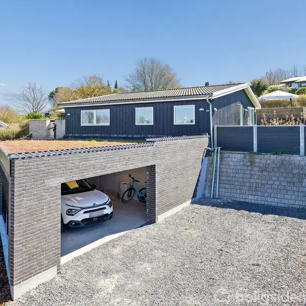 Garage med åben port indeholder en hvid bil og en cykel. Ovenpå ses et moderne hus med fladt tag omgivet af en grusbelagt indkørsel og grønne områder i et kvarter.